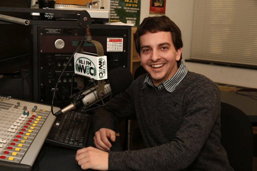 York College alumnus sits beside a WVYC 88.1 FM microphone and soundboard in the radio station studio.