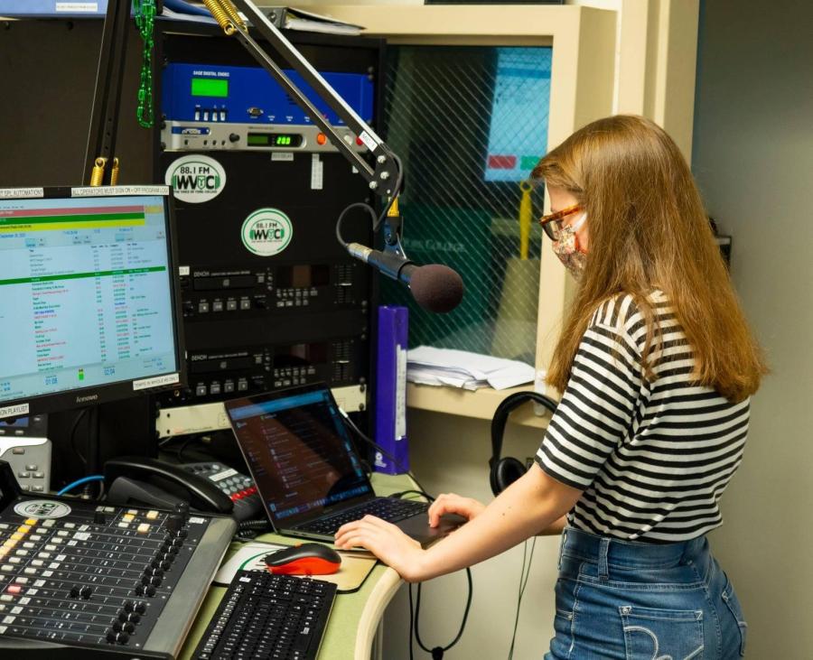 York College student works at a computer beside a microphone and broadcasting equipment in the WVYC radio studio.