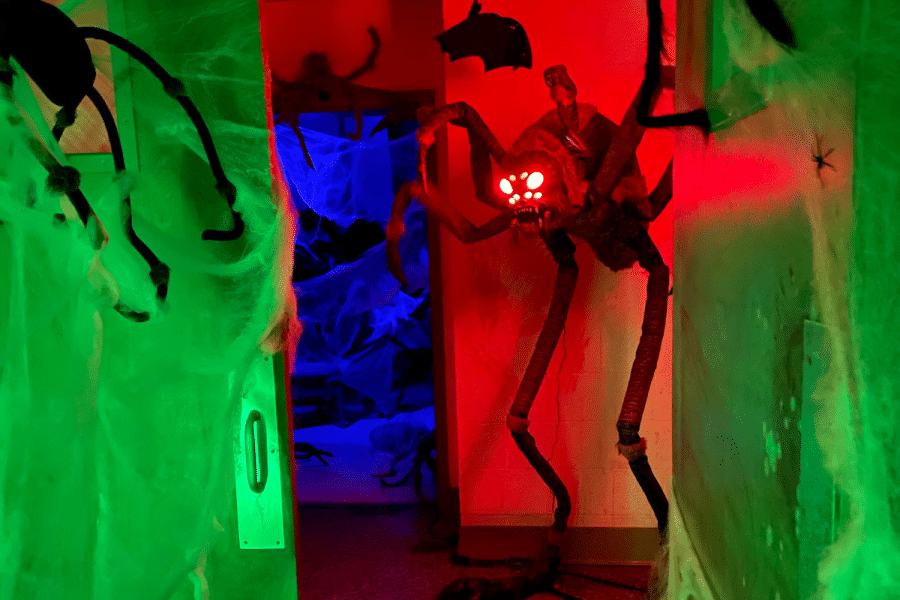 A Haunted House hallway glows with red, green, and blue lighting and features a large spider prop, cobwebs, bats, and other Halloween decorations.