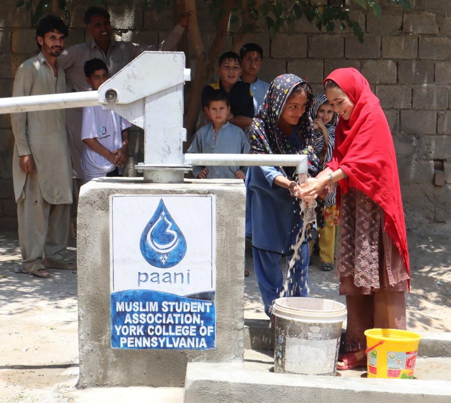 Children and adults gather around a newly constructed Paani Project well in Pakistan as two girls wash their hands with clean water.