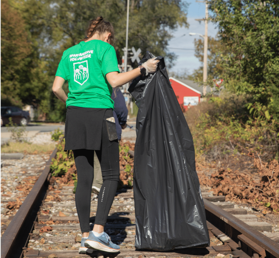 A SpartanServe volunteer wearing a green shirt collects litter along railroad tracks with a large trash bag.