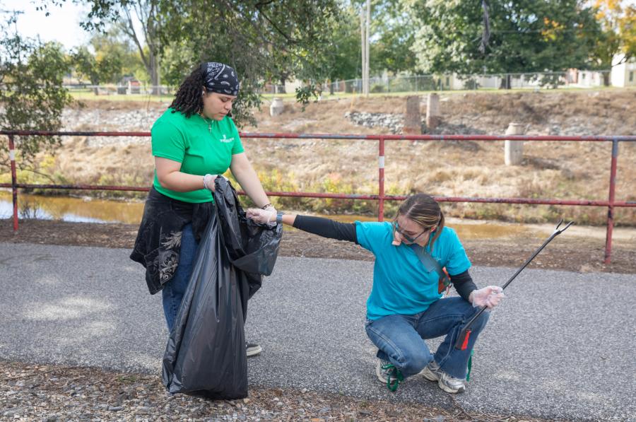 Two SpartanServe volunteers work together to collect litter near a walking path, using a trash bag and litter grabber.