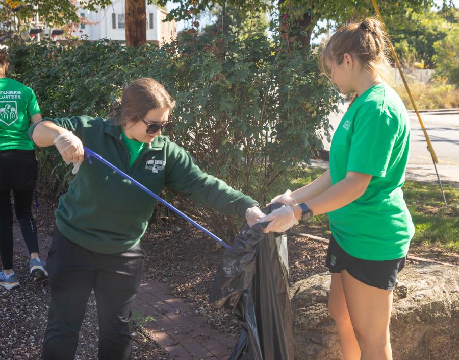 Two SpartanServe volunteers collect litter near a landscaped area, with one holding a trash bag and the other using a litter grabber.