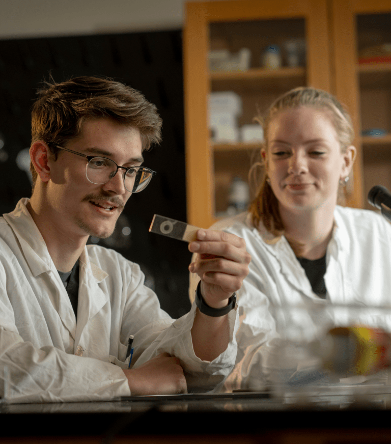 Two students in lab coats examine a microscope slide in a science lab.