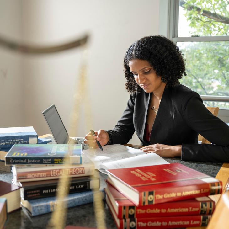 A student sits at a library table covered with law and criminology textbooks. The scales of justice sit on the desk, visible in the foreground.
