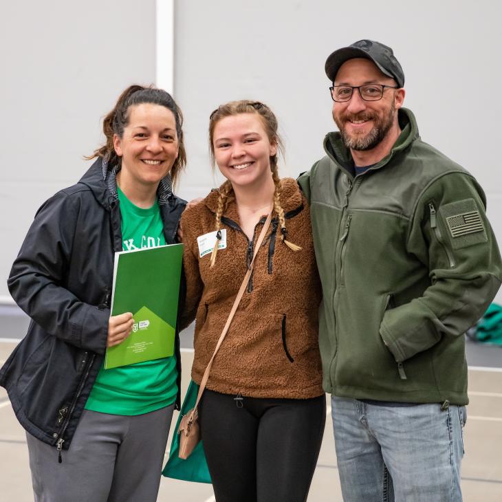 A student stands between her parents, smiling for a photo in the gymnasium.