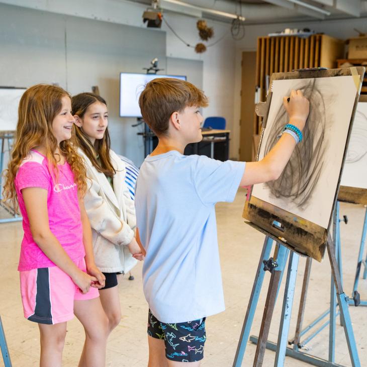 Three students stand facing an art project with one student drawing