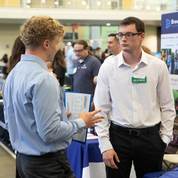Two people engaged in discussion at a professional networking event. One is wearing a green name tag indicating Alumni status.