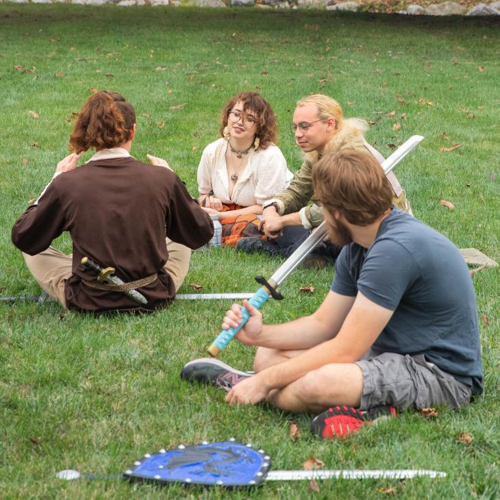 A group of students sitting and laughing together in a grassy field after a LARPing match.