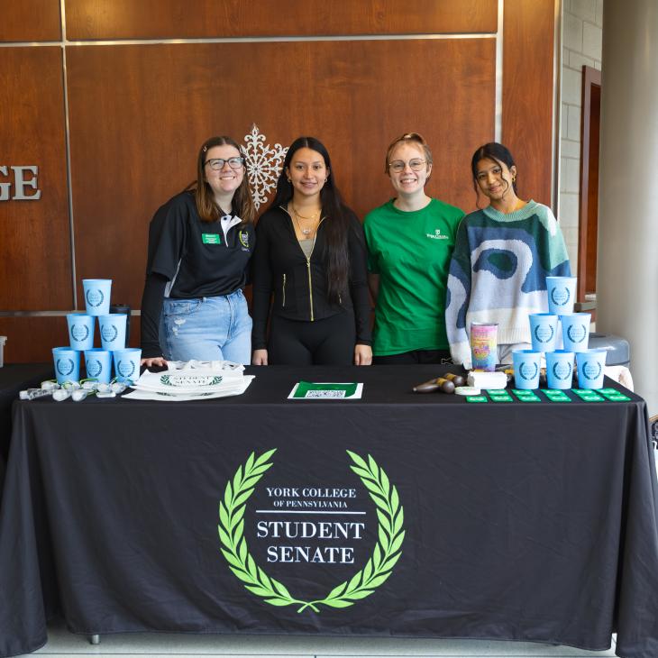 A group of students smile behind a table with a black "student senate" tablecloth.