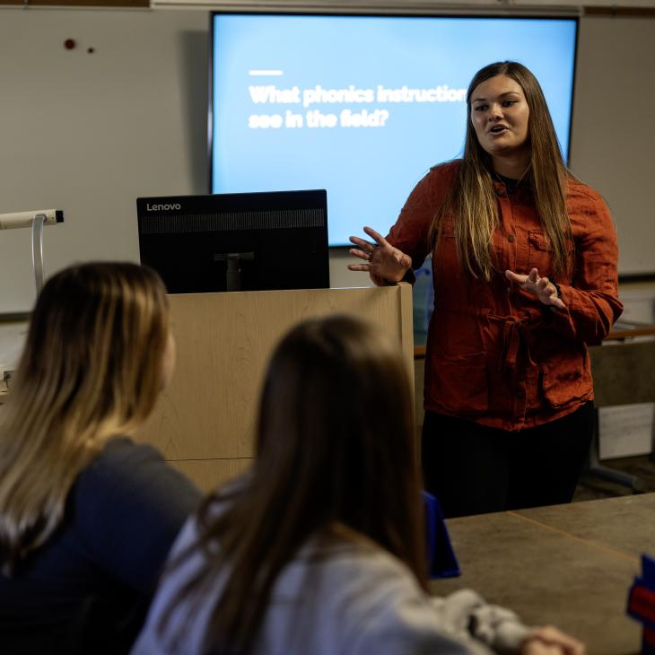 Instructor lecturing to a class with a projection screen visible in the background.