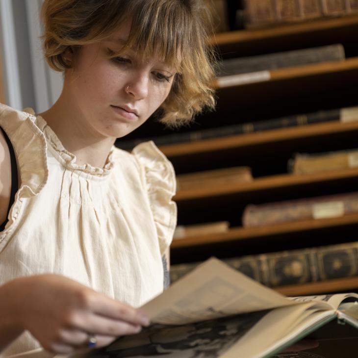 Person wearing a white shirt reading in a library, there is a tattoo visible on the person's arm and a bookshelf behind them.