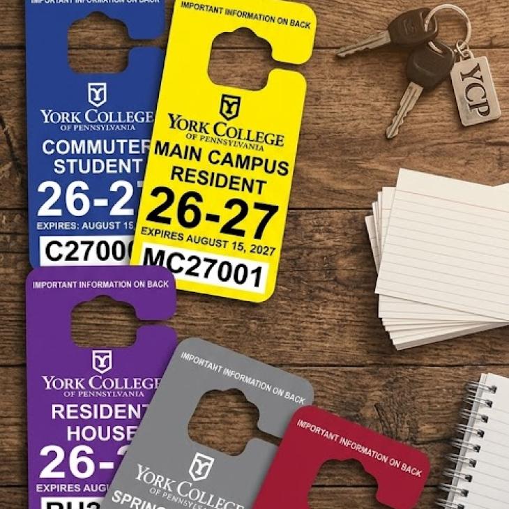 A collection of five York College of Pennsylvania parking permits in various colors (blue, yellow, purple, grey, and maroon) for the 2026-27 academic year, arranged on a wooden desk alongside keys and a notebook.
