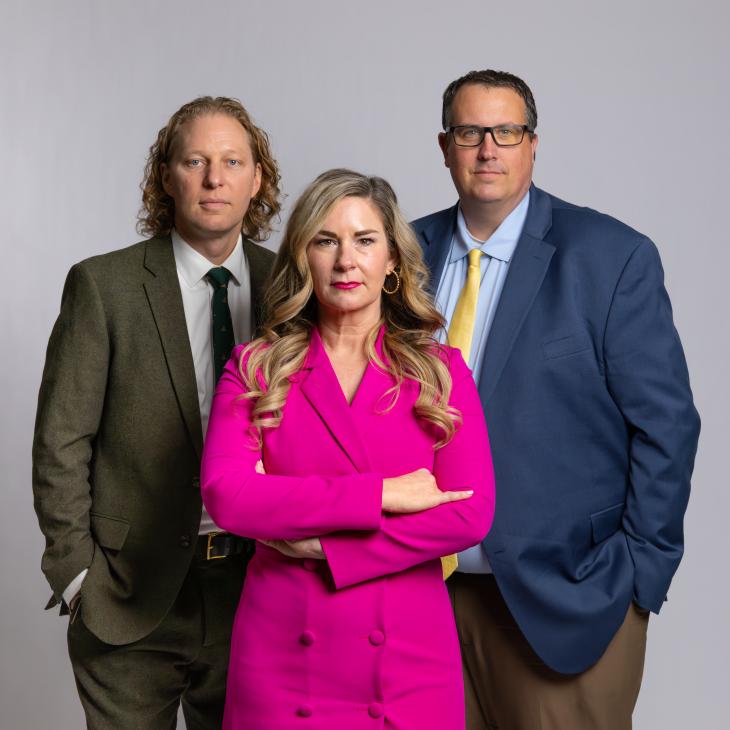 Three people dressed in professional clothes stand together against a grey backdrop.