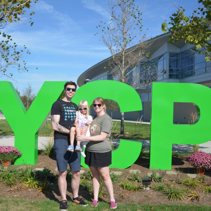 Family of 3 posing in front of the large, green YCP on west campus