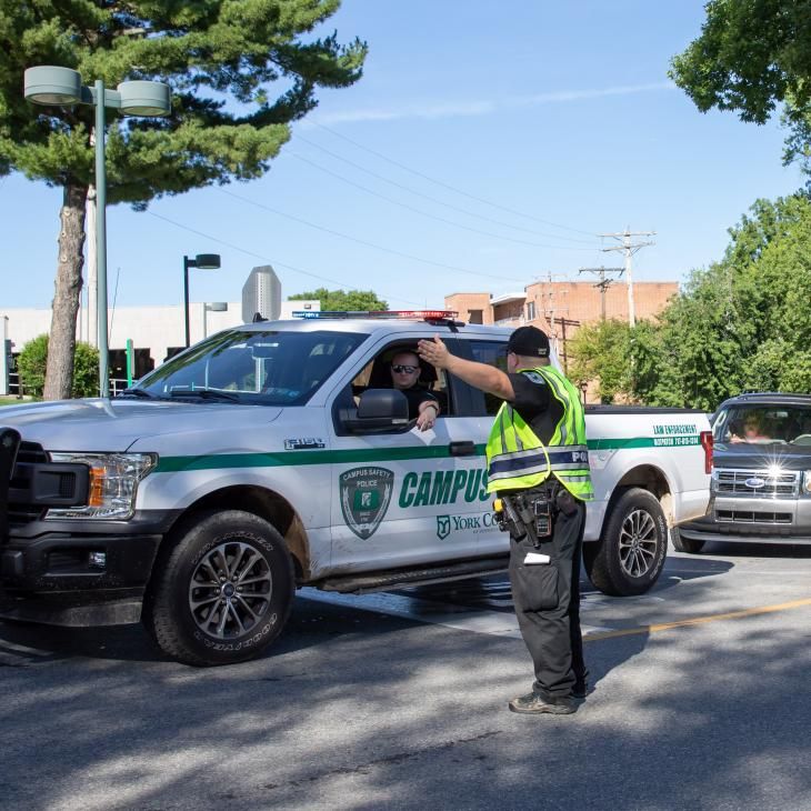 Campus safety officer directing traffic beside a campus police truck at a road closure with barricades on a sunny day.