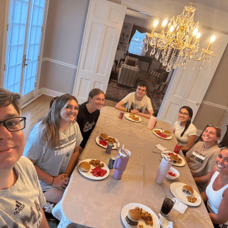 A group of students sits around a dining table together, smiling with plates of food in front of them.