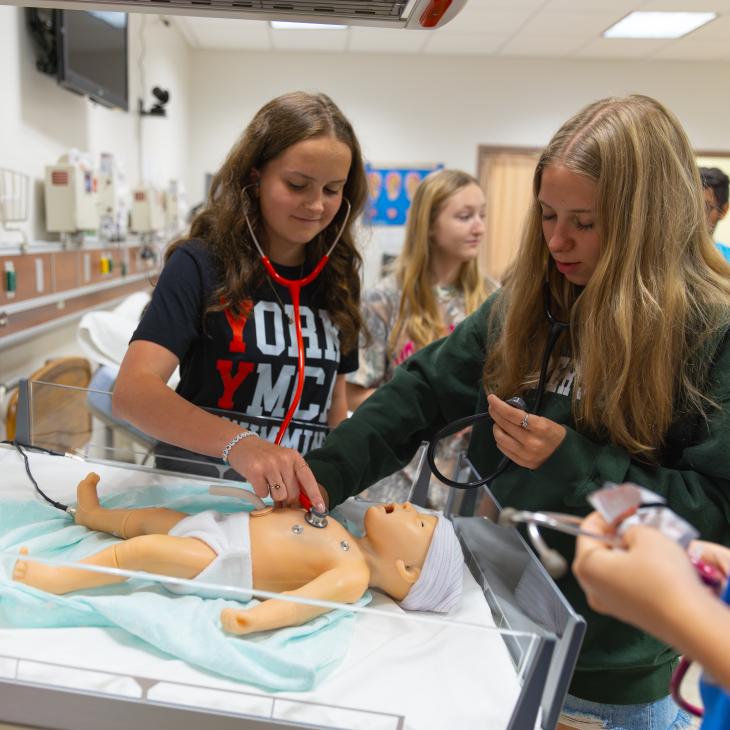 Students practice using stethoscopes on a medical training mannequin in a clinical lab.
