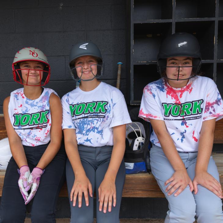 Three softball players wearing helmets and York t-shirts in the dugout.