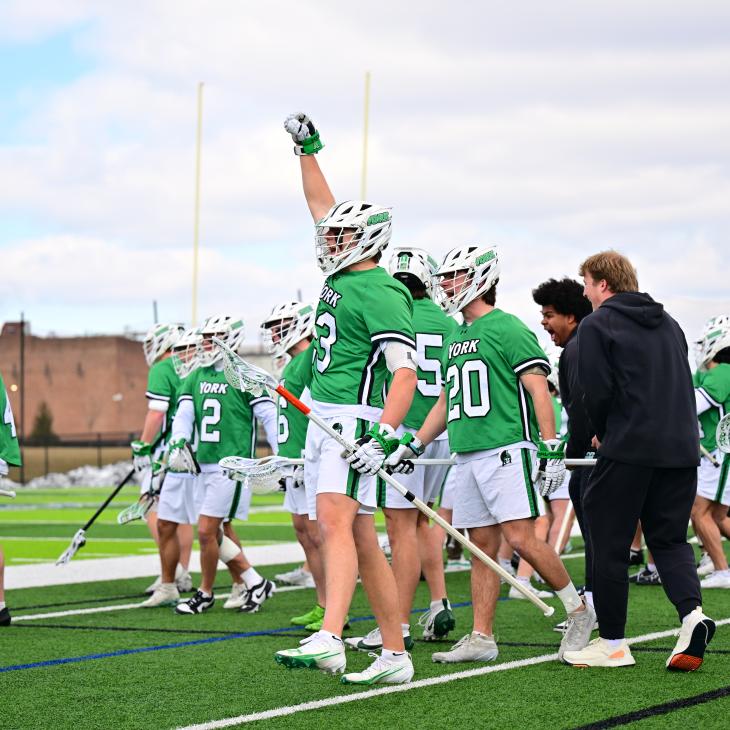 Lacrosse players on a field in their equipment with one player raising his hand.