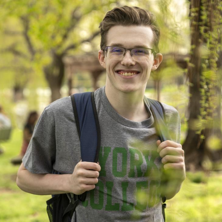 A student wearing a York College t-shirt smiles holding his backpack surrounded by willow trees.